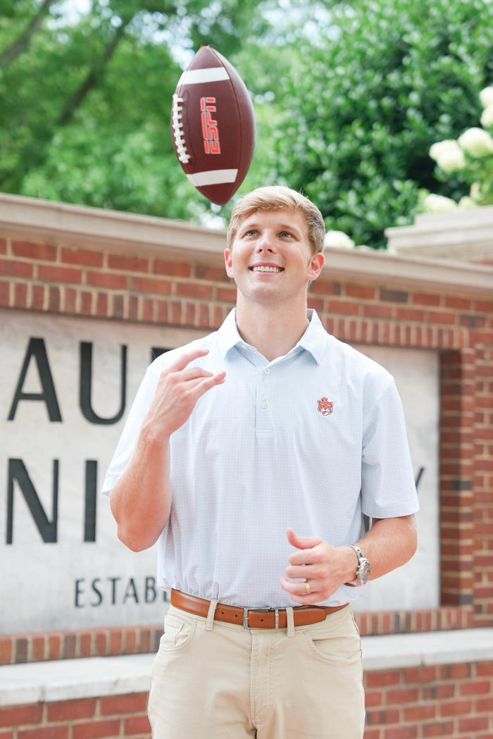 A young man smiling and looking up at a football balanced on his finger, wearing a light blue polo shirt with a small crimson logo on the chest, khaki pants, and a brown belt. He is standing outdoors in front of a brick wall with a sign and green trees in the background.
