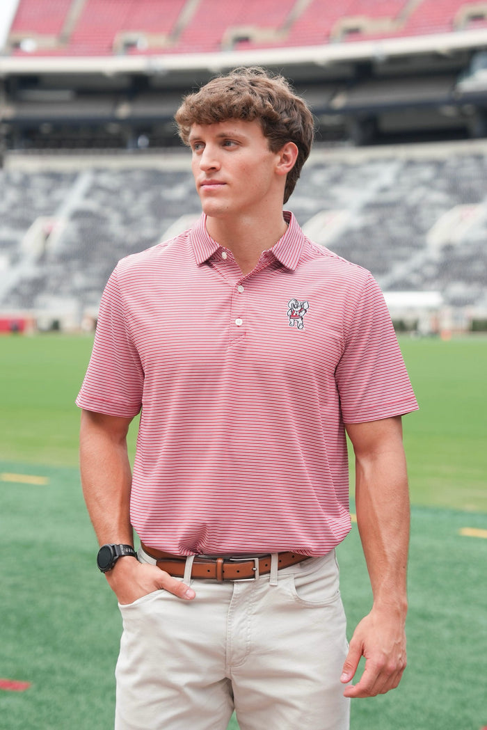 A man wearing a pink and white striped polo shirt with a small embroidered logo on the chest, khaki pants, and a black watch, standing on a green sports field with stadium seating in the background.