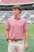 A man wearing a pink and white striped polo shirt with a small embroidered logo on the chest, khaki pants, and a black watch, standing on a green sports field with stadium seating in the background.