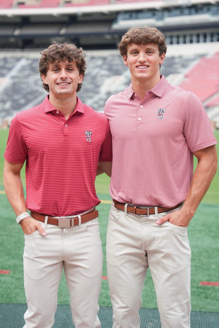 Two young men standing side by side in a stadium, wearing matching polo shirts. The man on the left wears a red and white striped polo shirt with khaki pants, and the man on the right wears a pink and white striped polo shirt with khaki pants.