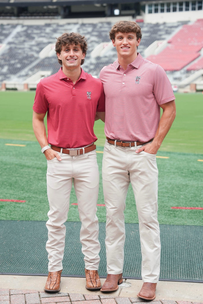 Two young men standing on a sports field wearing matching polo shirts and khaki pants. The man on the left wears a coral-red polo shirt, and the man on the right wears a pink polo shirt.