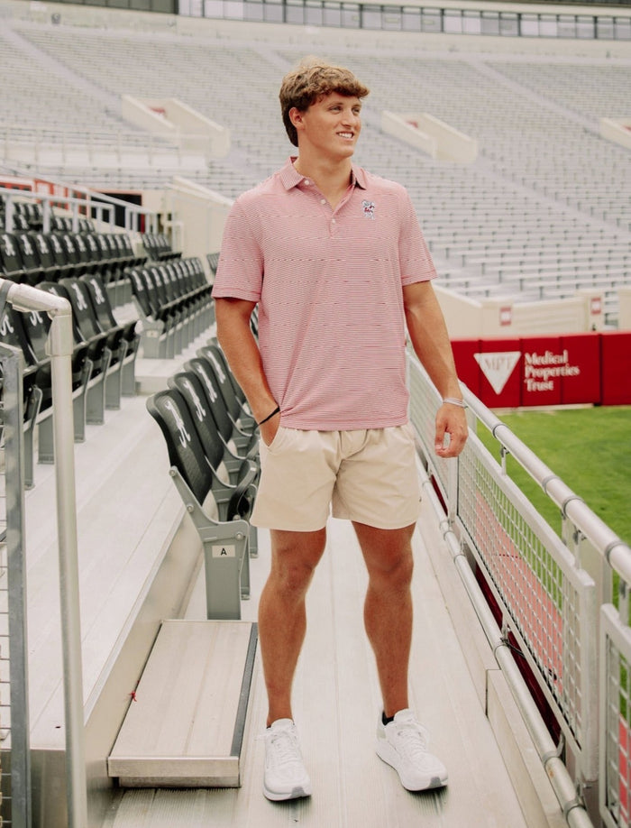 A man wearing a pink polo shirt with elephant embroidery and khaki shorts standing on a stadium concourse.