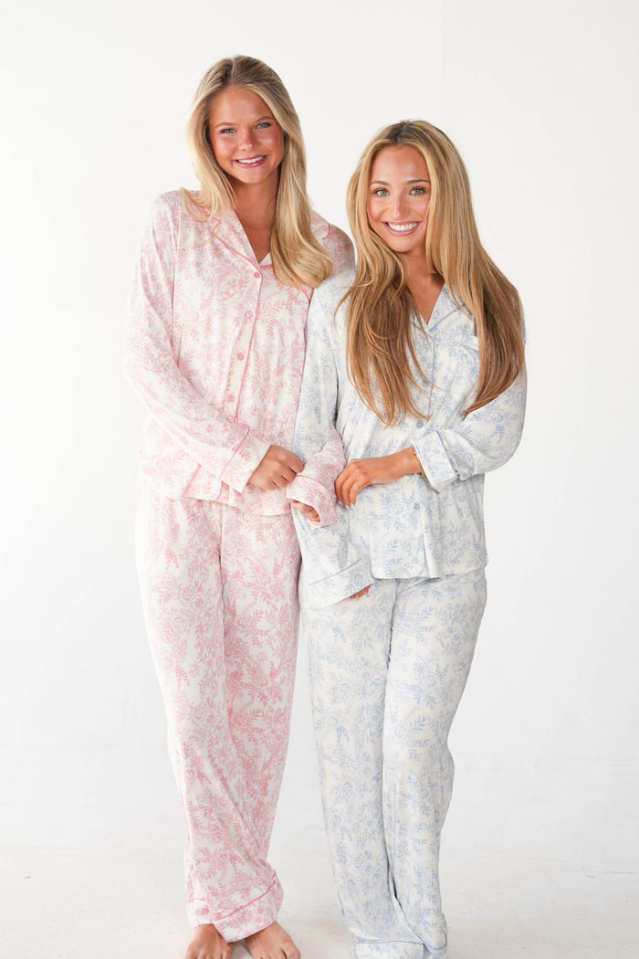 Two women modeling matching long-sleeve pajama sets against a plain white studio background. The woman on the left wears a light pink pajama set with a subtle floral pattern, and the woman on the right wears a light blue pajama set with a matching floral print.