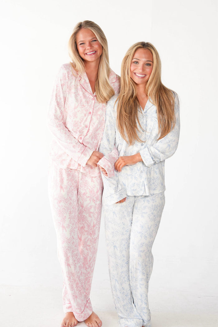 Two women standing side by side wearing matching floral pajama sets, one in light pink and one in light blue, against a plain white studio background.