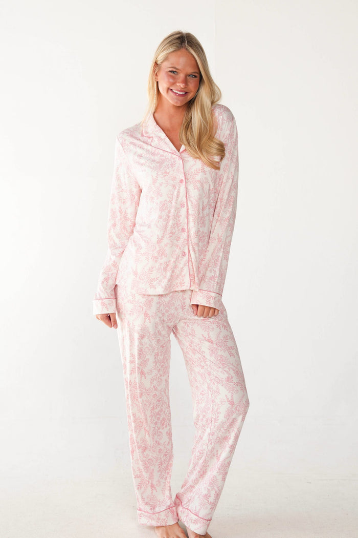 A woman modeling a light pink and white floral print pajama set, consisting of a long-sleeve button-up shirt and matching pants, against a plain white studio background.