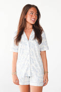 A woman modeling a matching short-sleeve button-front top and shorts pajama set in a light blue and white floral toile pattern, set against a plain white studio background.