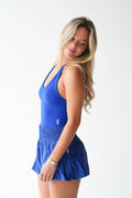 A woman modeling a bright blue athletic two-piece set consisting of a scoop-neck tank top and a matching high-waisted ruffled mini skirt, set against a plain white studio background.