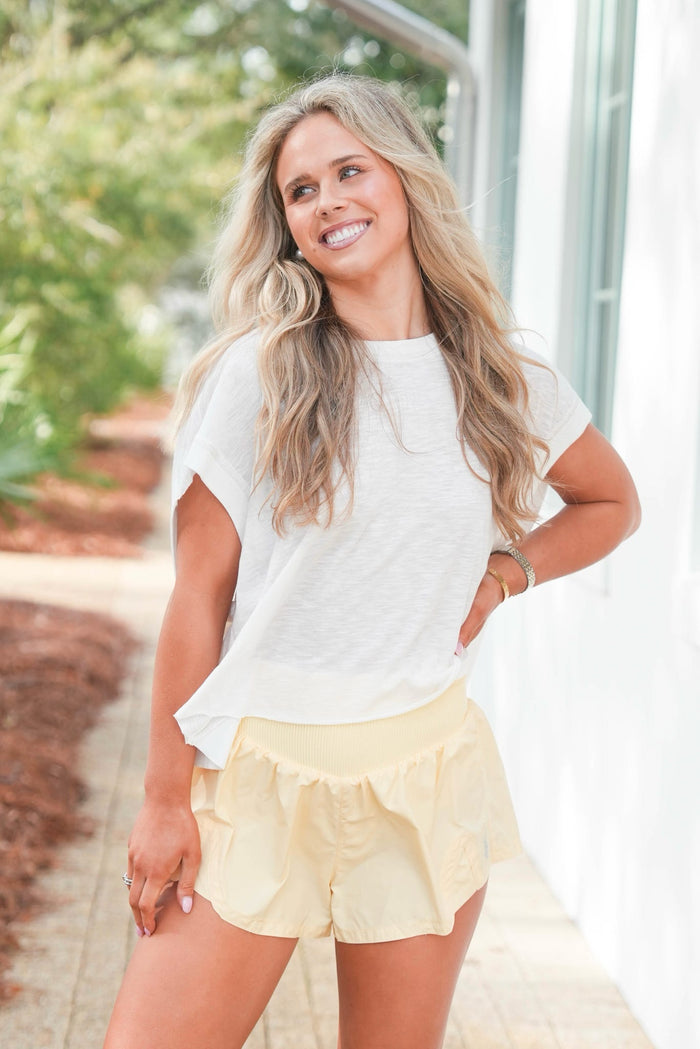 Woman wearing a white top and yellow shorts standing outdoors with greenery in the background
