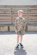 Young boy in camouflage shirt and shorts standing outdoors with wooden slats in the background