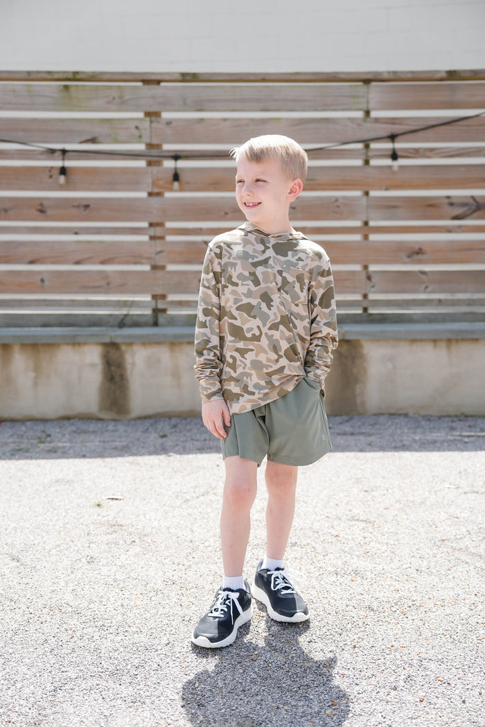 Young boy in camouflage shirt and shorts standing outdoors with wooden slats in the background