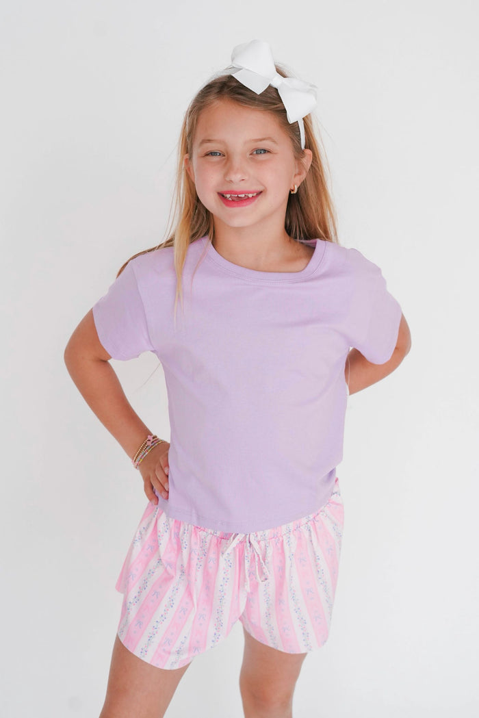 Young girl wearing a purple top and pink shorts with white stripes on a white background