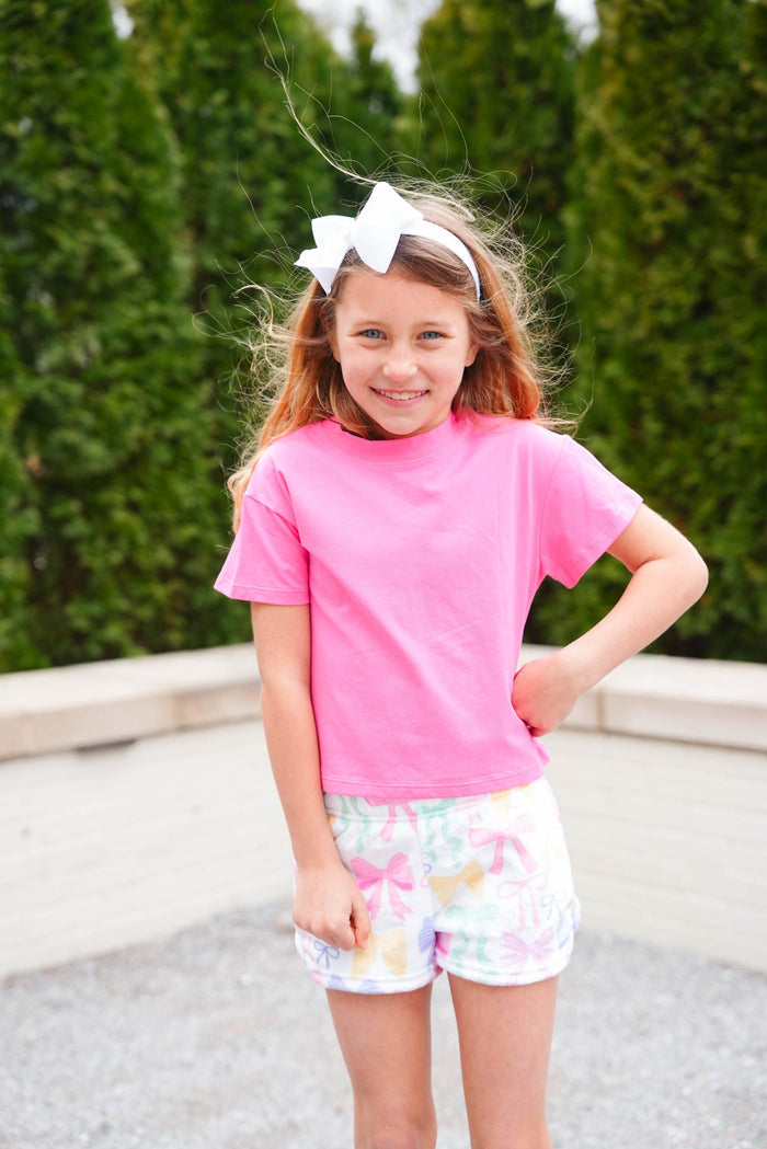 Young girl wearing a pink shirt and patterned shorts with a white bow in her hair, standing outdoors.