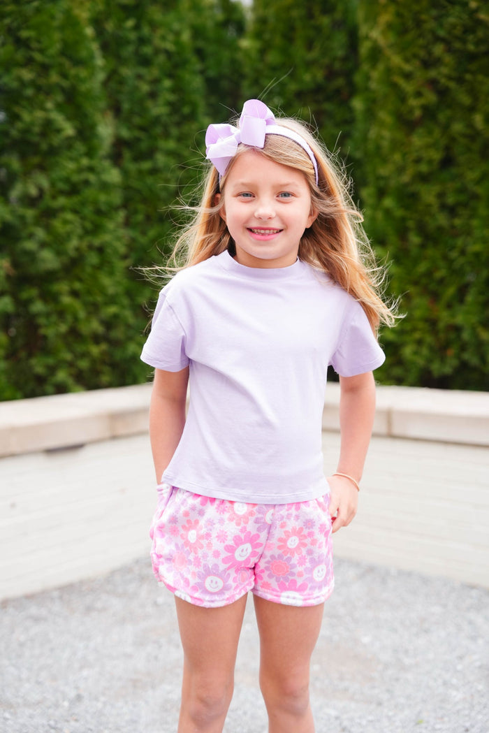 Young girl wearing a purple top and pink and purple floral shorts is posing outside.