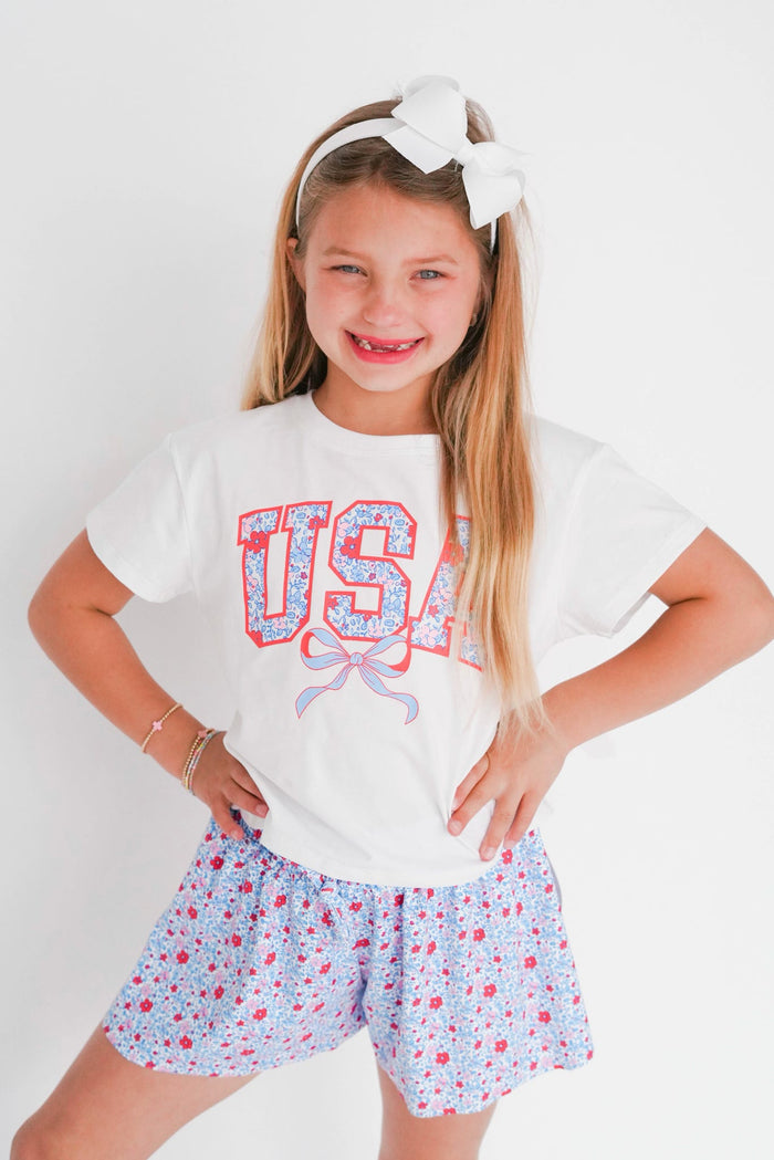 Young girl wearing a 'USA' t-shirt with floral design and shorts on a white background