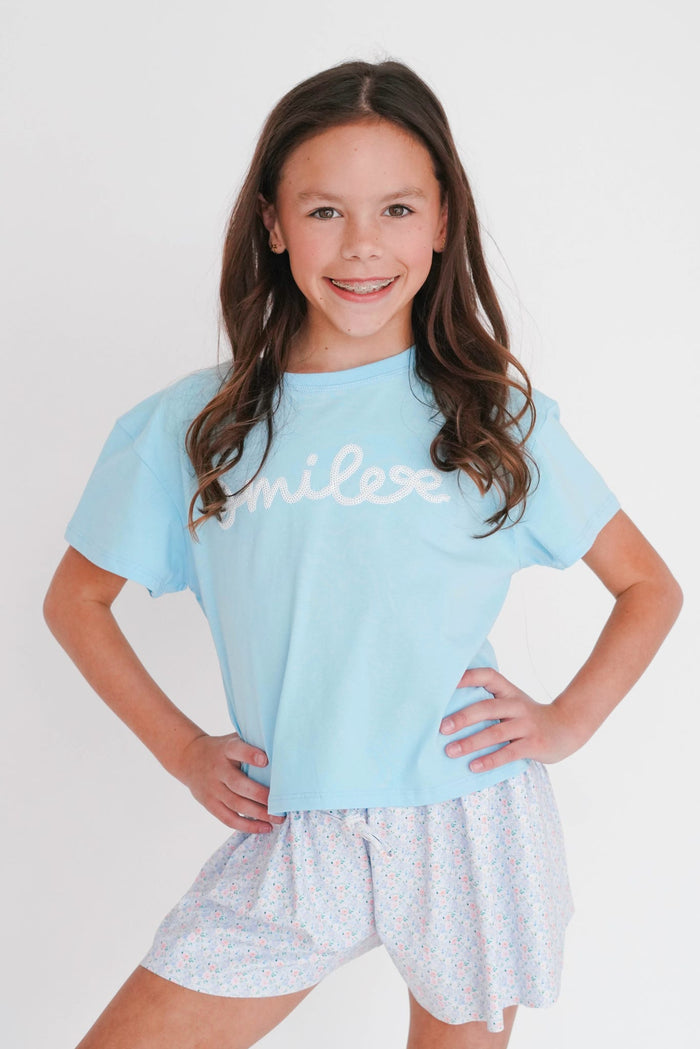 Young girl wearing a light blue t-shirt with 'smile' and white shorts on a plain background
