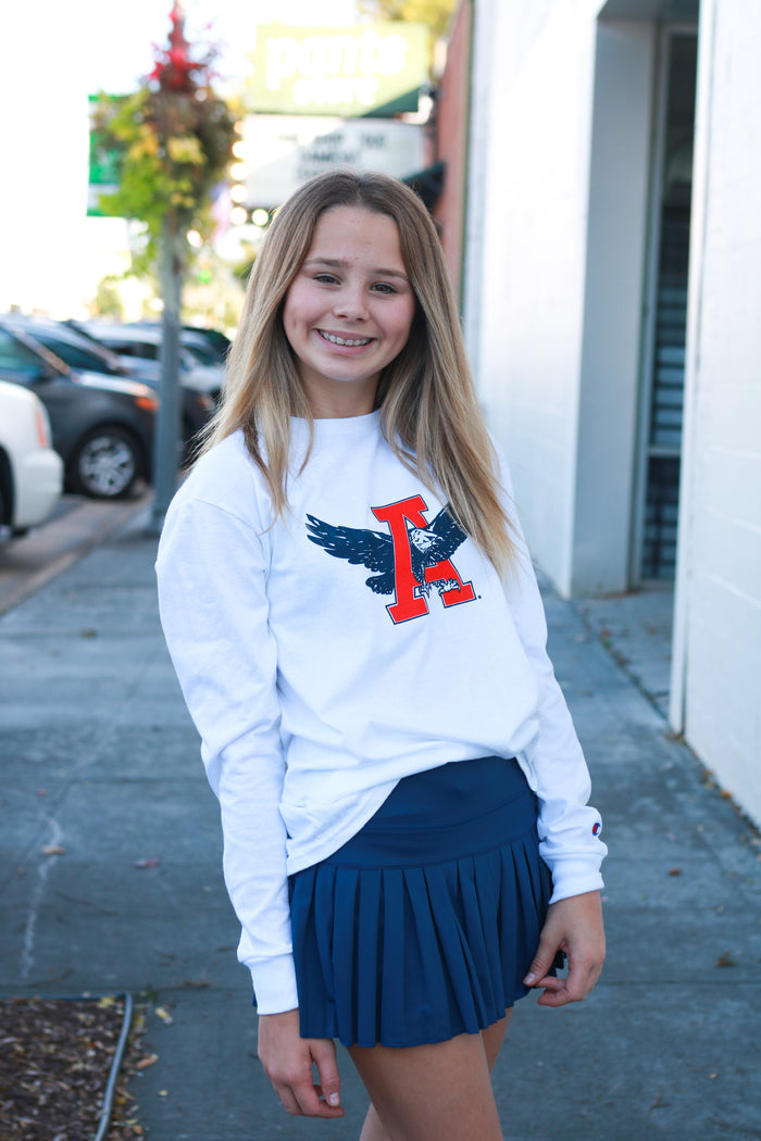 Young girl wearing a white sweatshirt with a logo and blue skirt standing on a sidewalk.
