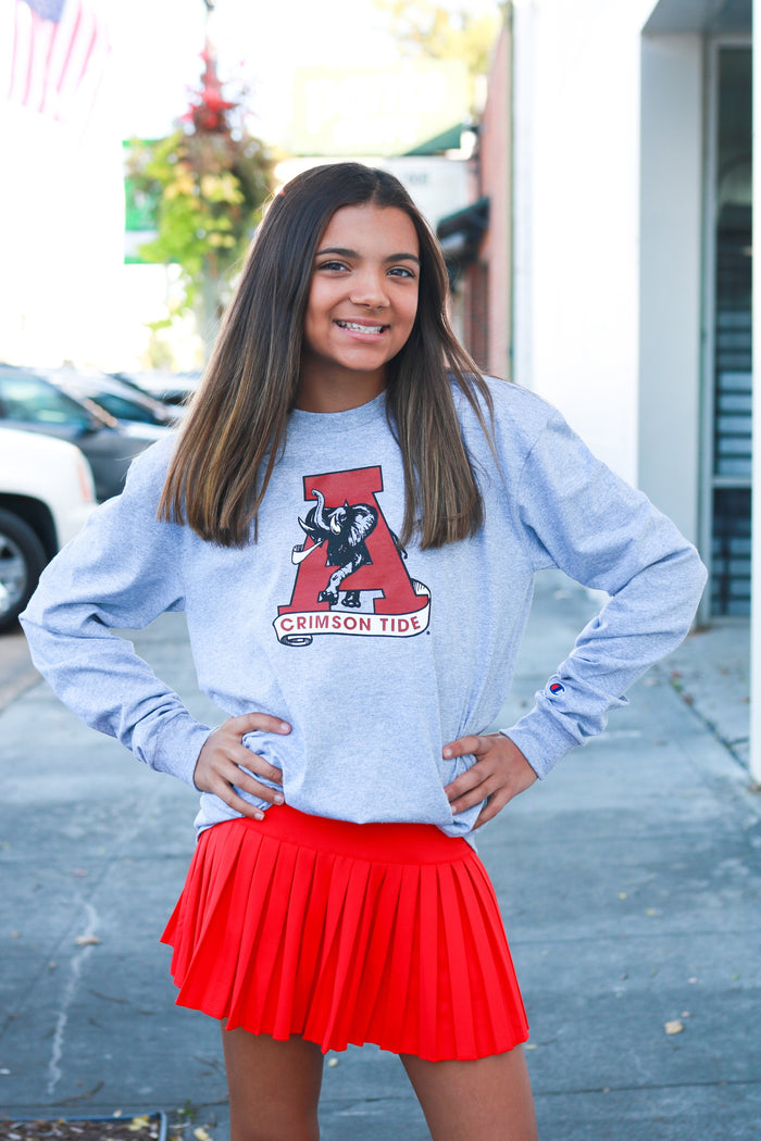 Girl wearing a gray sweatshirt with a logo and a red skirt standing on a sidewalk.