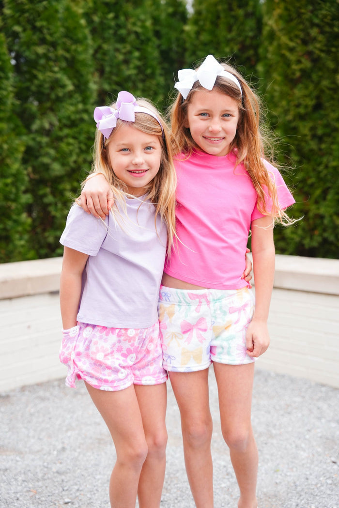 Two young girls standing close to each other, wearing hair bows on headbands. One girl is in a purple shirt and the other in a pink shirt, both with floral shorts.
