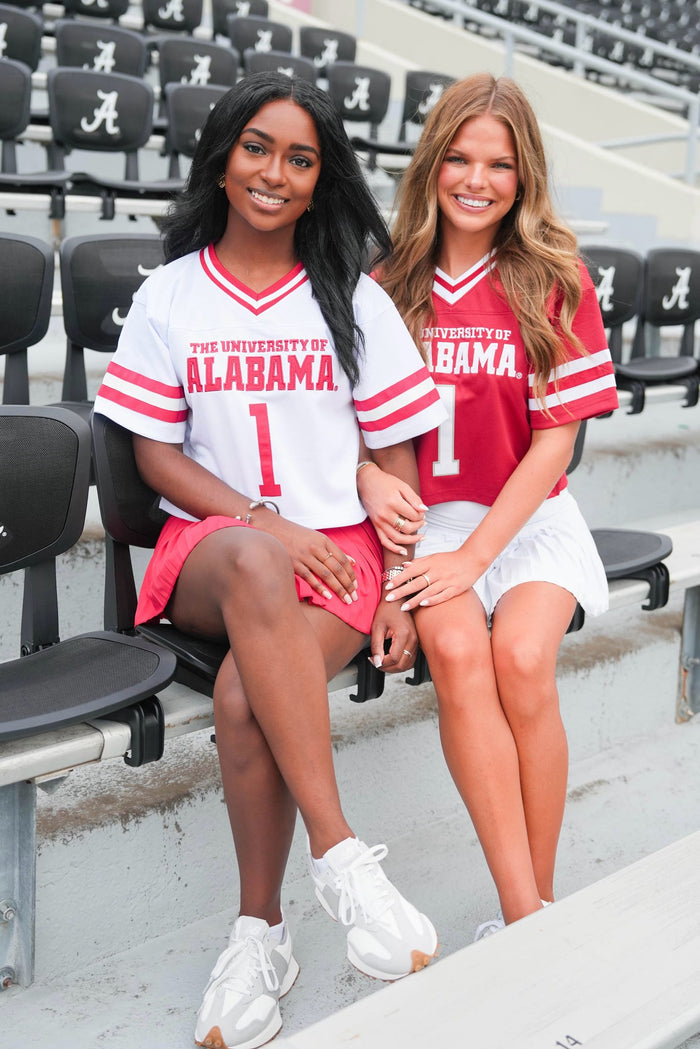 Two women wearing University of Alabama jerseys sitting on a bench in a stadium.