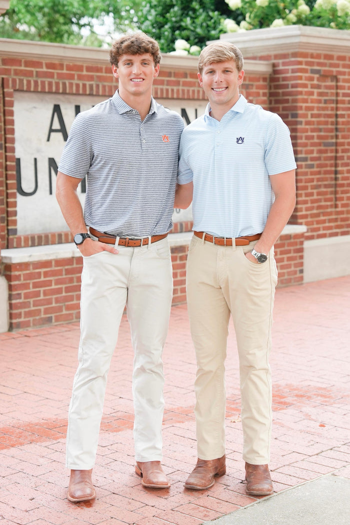 Two young men standing outdoors in front of a brick wall and brick pavement, wearing polo shirts and khaki pants. The man on the left wears a gray and white striped polo shirt, and the man on the right wears a light blue polo shirt.