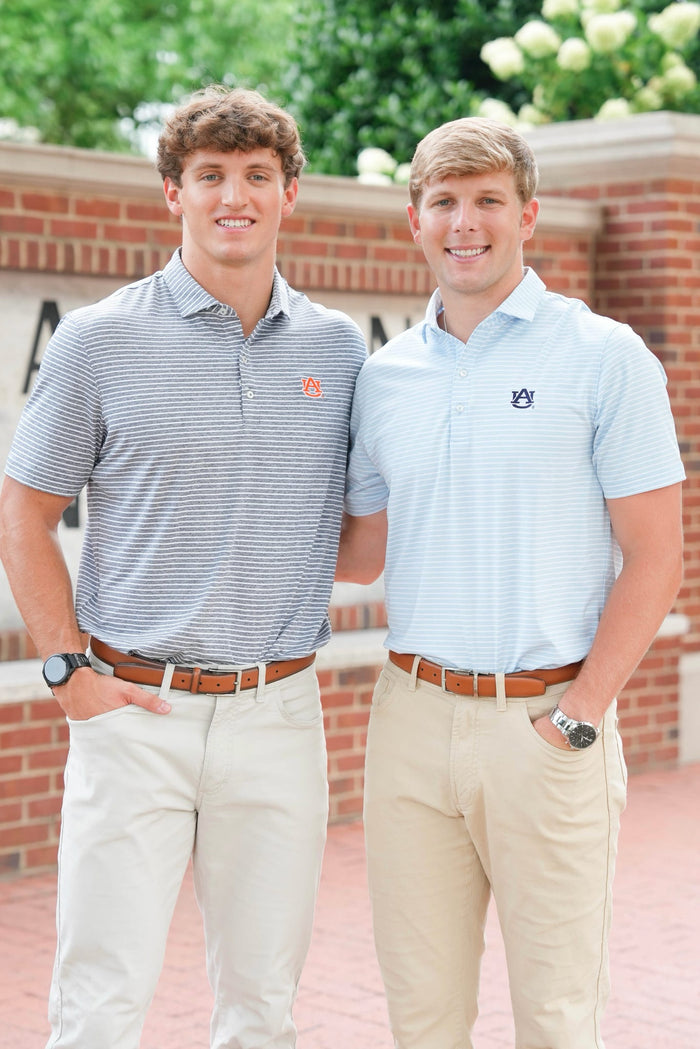 Two young men standing outdoors in front of a brick wall and greenery, each wearing a short-sleeve polo shirt and khaki pants. The shirt on the left has thin gray stripes with a small orange logo, and the shirt on the right is light blue with a small dark logo.