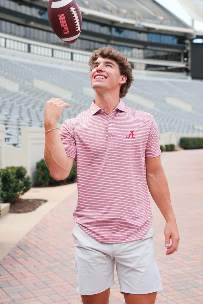 Man in a red striped polo shirt and light shorts holding a football in front of a stadium.