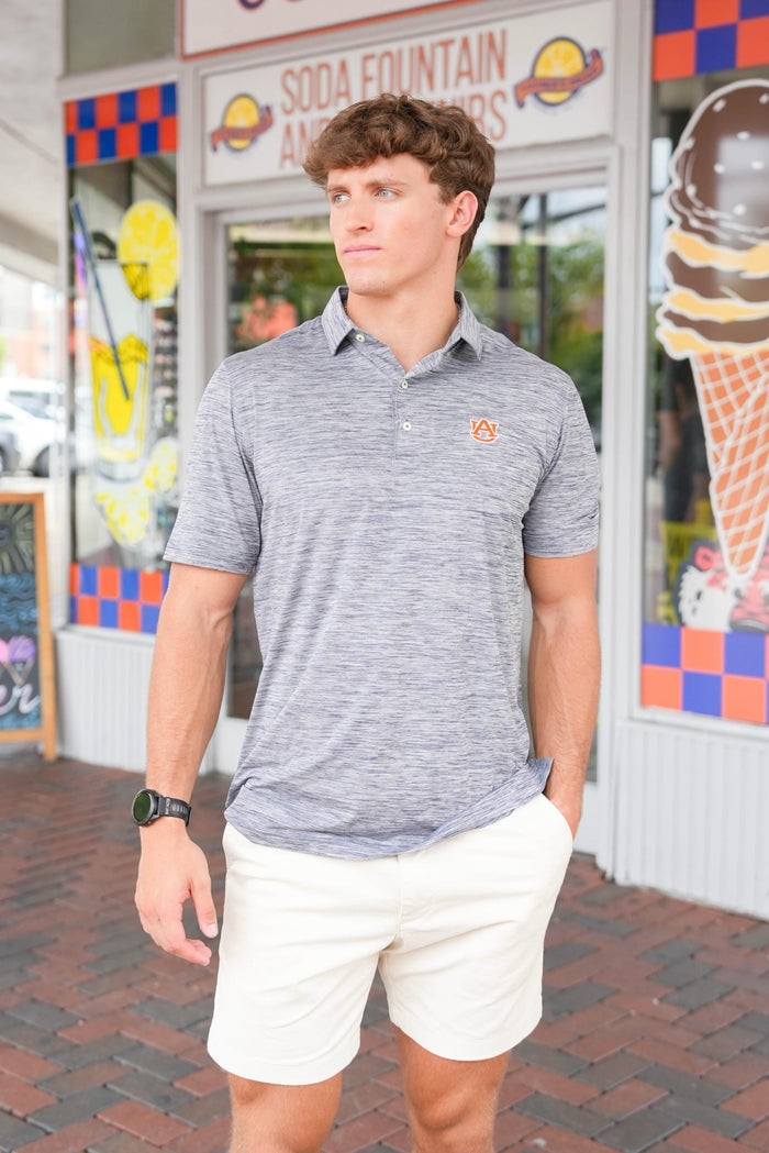A man wearing a heather gray short-sleeve polo shirt with a small orange logo on the chest and off-white shorts, standing in front of a colorful storefront.