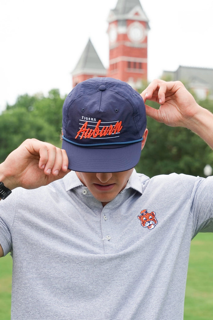 A man wearing a navy blue baseball cap with 'Tigers Athletics' embroidered in orange and white, holding the brim over his eyes. He is wearing a gray polo shirt with a matching tiger logo on the chest, set against a blurred background of a red brick clock tower and green trees.