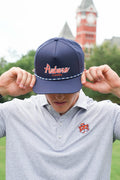 A man wearing a navy blue Auburn Tigers baseball cap with 'Auburn Tigers' embroidered on the front, holding the brim down over his eyes. He is wearing a gray polo shirt with an Auburn Tigers logo on the chest, with a blurred clock tower in the background.
