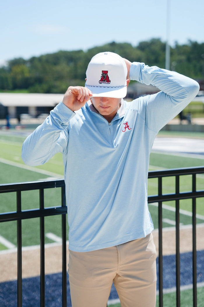 A person wearing a light blue long-sleeved shirt and beige pants is adjusting a white rope hat with an Alabama logo.