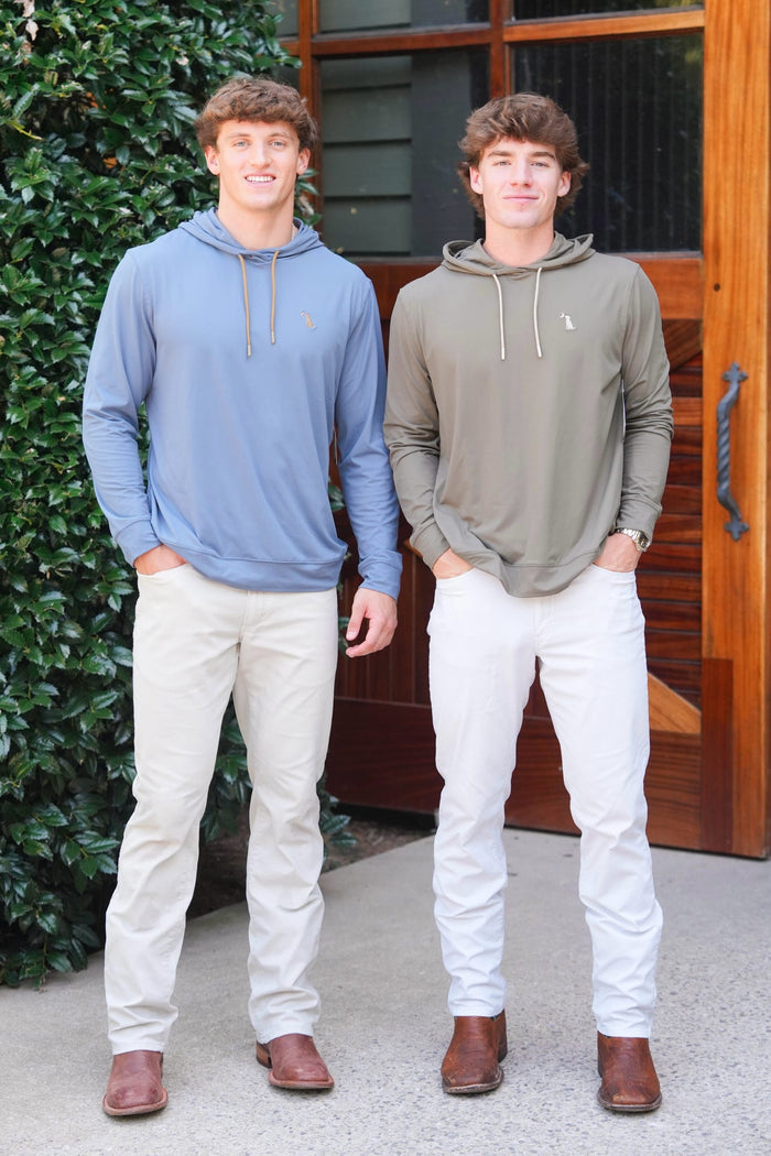 Two young men standing outdoors in front of a wooden door and green hedge, wearing Polo Ralph Lauren hoodies. The man on the left wears a light blue hoodie with beige pants, and the man on the right wears an olive green hoodie with white pants.