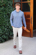 A man wearing a light blue long-sleeve hoodie with a small embroidered polo player logo on the chest, paired with beige pants and brown shoes, standing in front of a wooden door and green bushes.