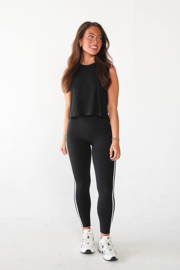 A woman modeling a black sleeveless crop top and black leggings with white side stripes, paired with white sneakers, against a plain white studio background.