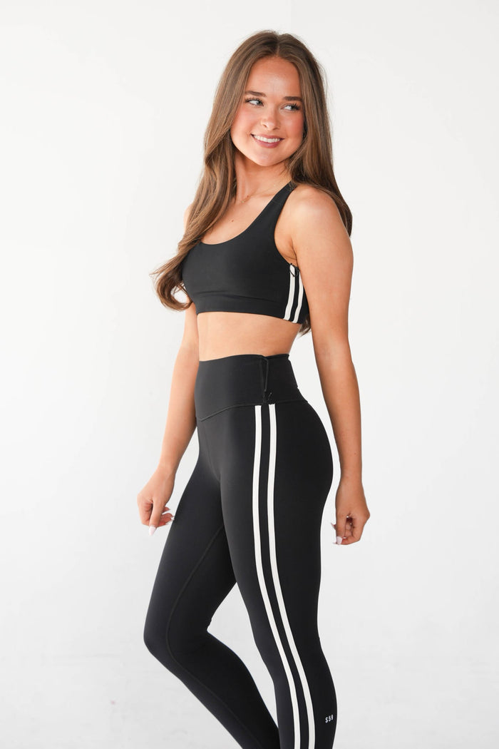 A woman modeling a matching black activewear set with white double stripes, consisting of a sports bra and high-waisted leggings, against a plain white studio background.