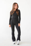 A woman modeling a black long-sleeve athletic top and matching black flared leggings with white sneakers, standing against a plain white studio background.