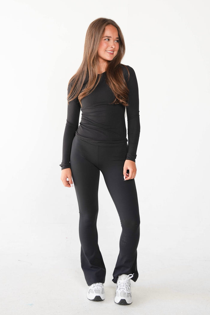 A woman modeling a black long-sleeve athletic top and matching black flared leggings with white sneakers, standing against a plain white studio background.