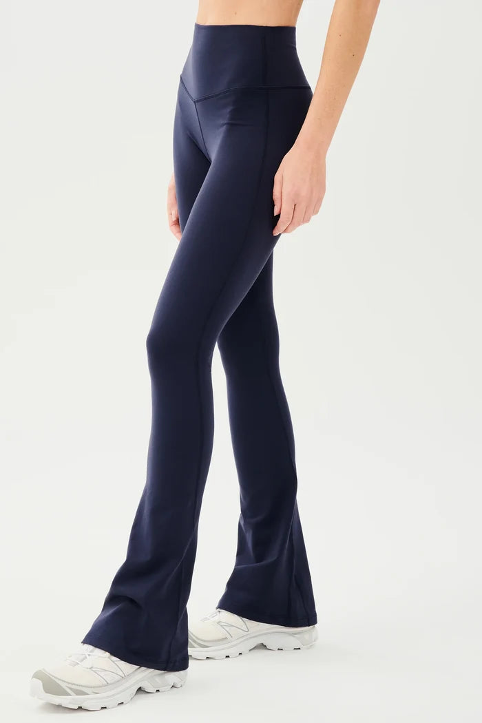 A pair of high-waisted black flare leg leggings, modeled in a standing position. The person is wearing sports shoes with blue accents.