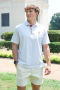A man wearing a white short-sleeve polo shirt with a red inner collar and a small crest logo on the chest, paired with light beige shorts, standing outdoors in front of a light-colored building and green hedges.