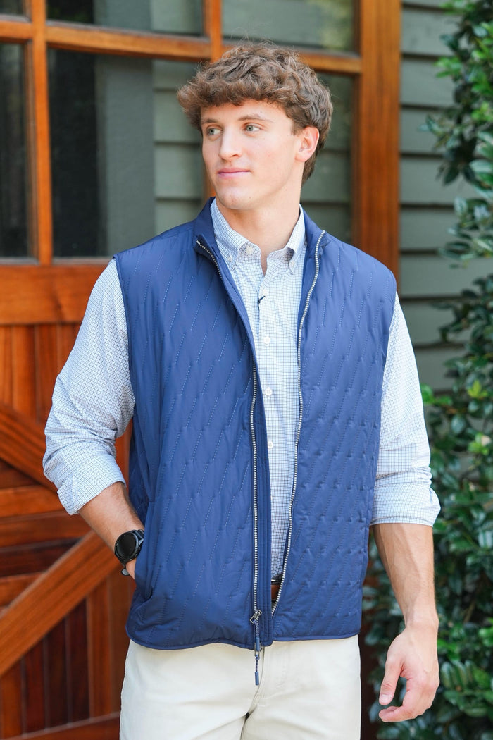 A man wearing a navy blue quilted puffer vest over a light blue and white checkered button-down shirt, paired with beige pants, standing in front of a wooden door and green foliage.