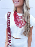 Woman wearing a white top and red necklace with 'Alabama' strap over shoulder against a light blue brick wall.