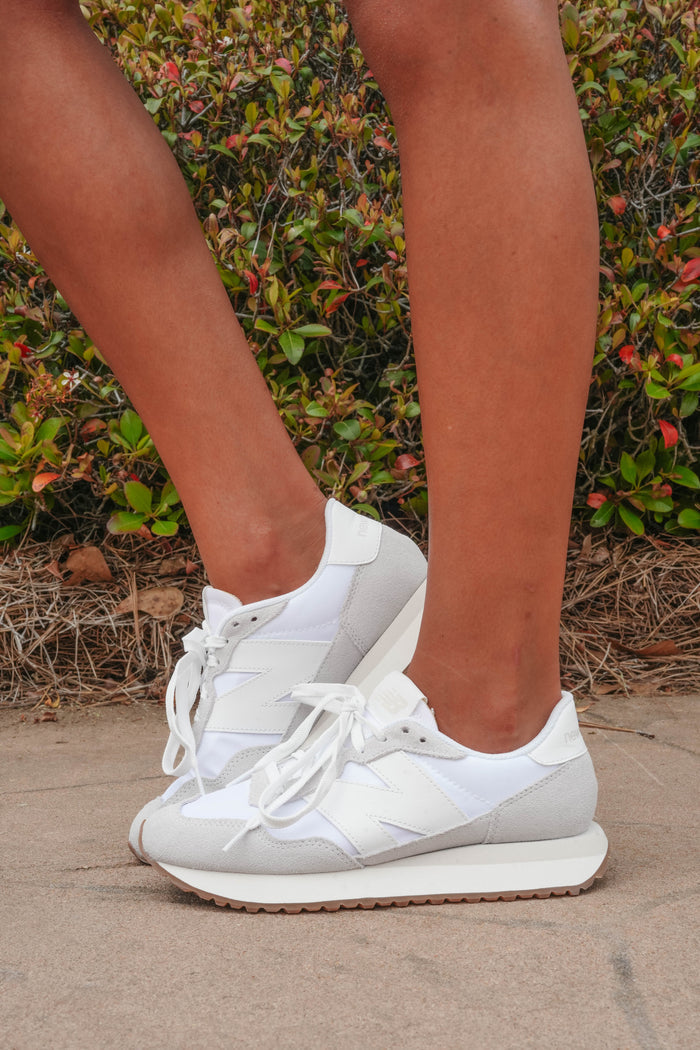 A pair of women's New Balance 237 white sneakers with a grey 'N' logo, shown from the side with the wearer's legs in the frame.