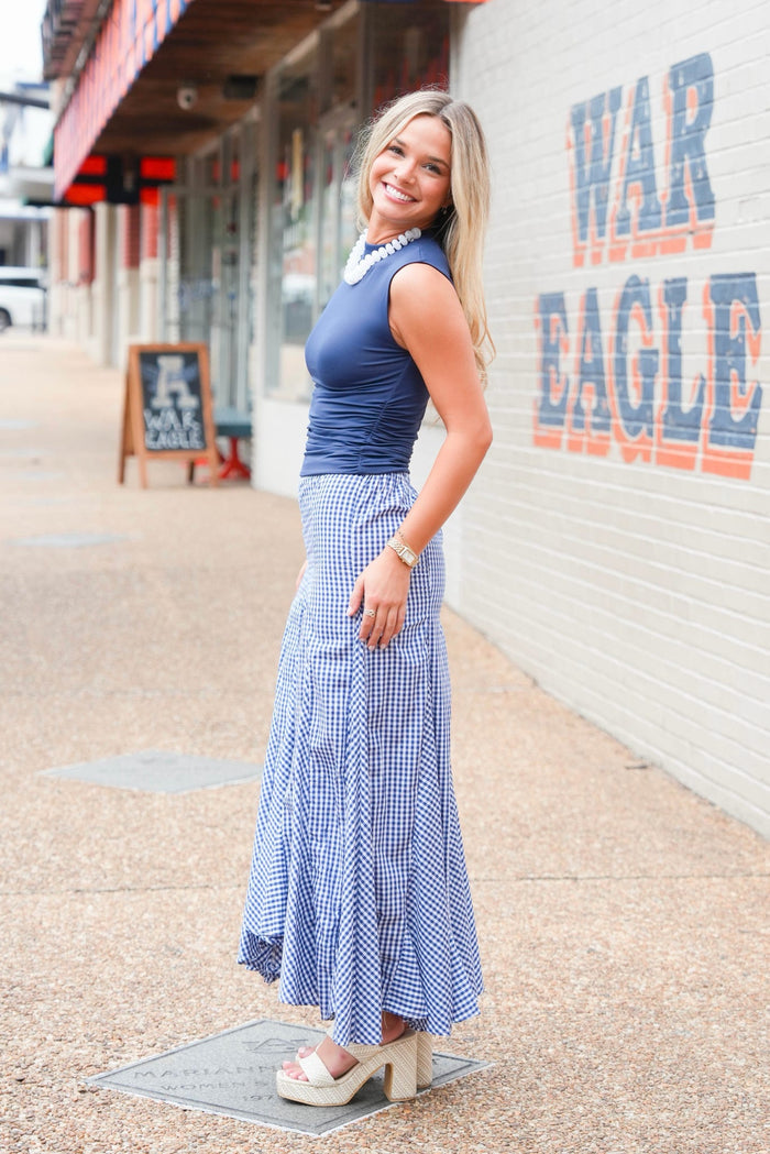 A woman modeling a sleeveless navy blue top and a matching blue-and-white gingham maxi skirt, standing on a sidewalk next to a white brick wall with 'WAR EAGLE' painted on it.