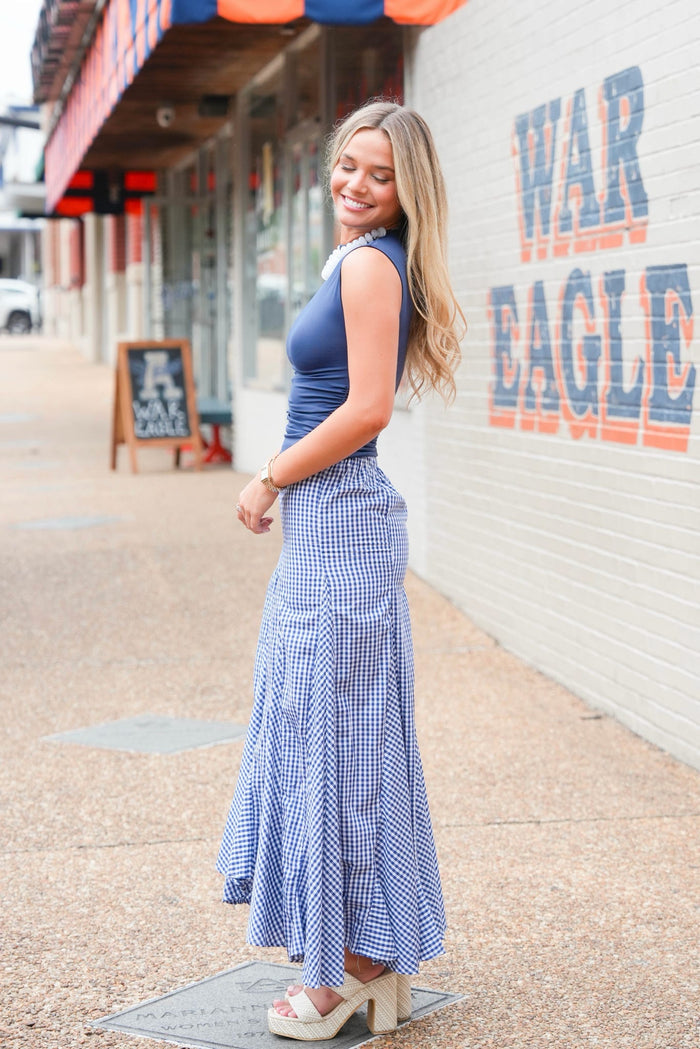 A woman stands outdoors on a sidewalk next to a white brick wall with 'WAR EAGLE' painted on it, wearing a sleeveless blue top and a long blue-and-white gingham maxi skirt with large patch pockets.
